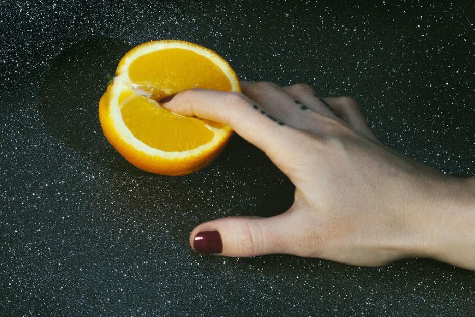 Eine Hand drückt einen Finger in eine halbe Orange vor einem dunklen, sternenartigen Hintergrund.