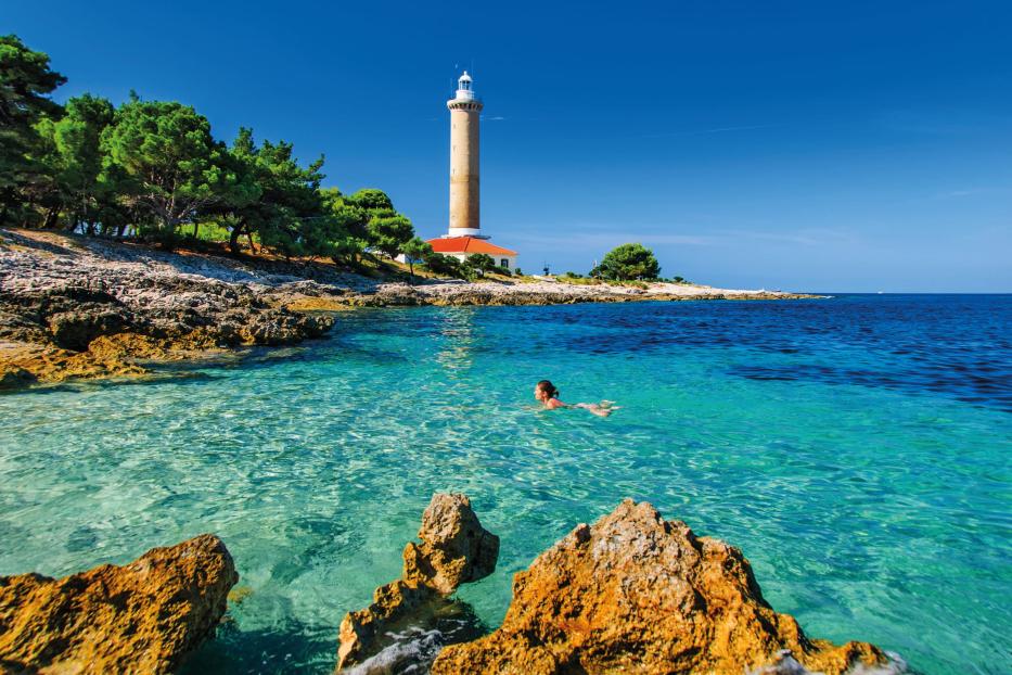 Eine Frau schwimmt im türkisfarbenen Wasser vor einem Leuchtturm.