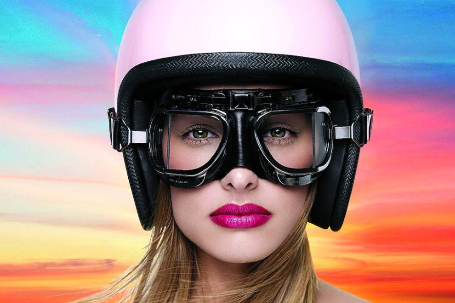 Eine Frau mit rosa Helm und Fliegerbrille vor einem farbenfrohen Himmel.