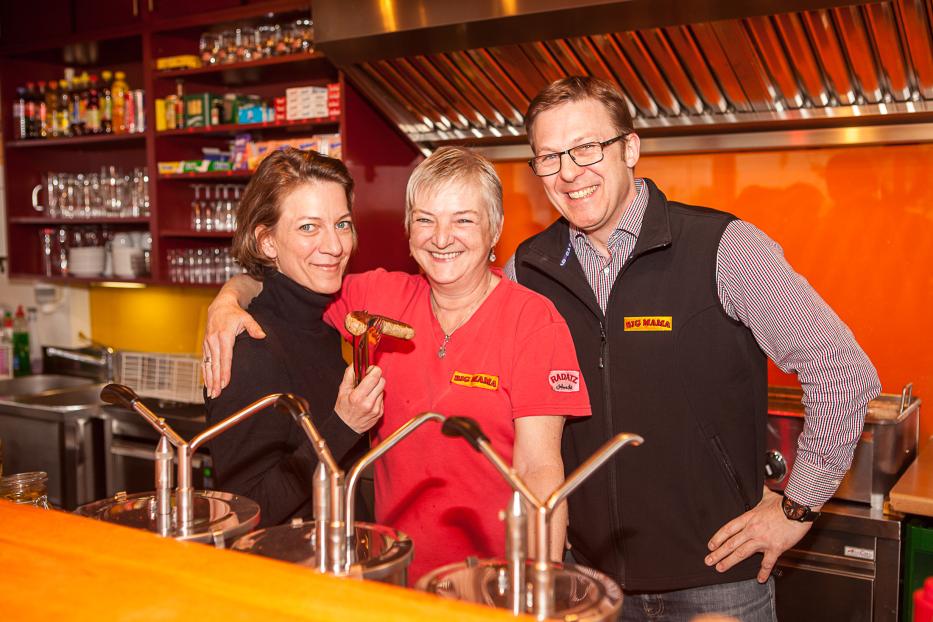 Drei Personen stehen lächelnd in einer Küche, eine hält eine Wurst in der Hand.