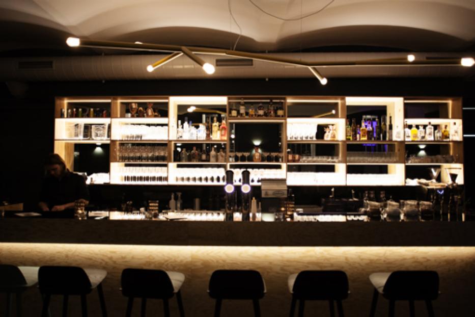 Eine Bar mit beleuchteten Regalen voller Flaschen und Gläser sowie Barhockern davor.