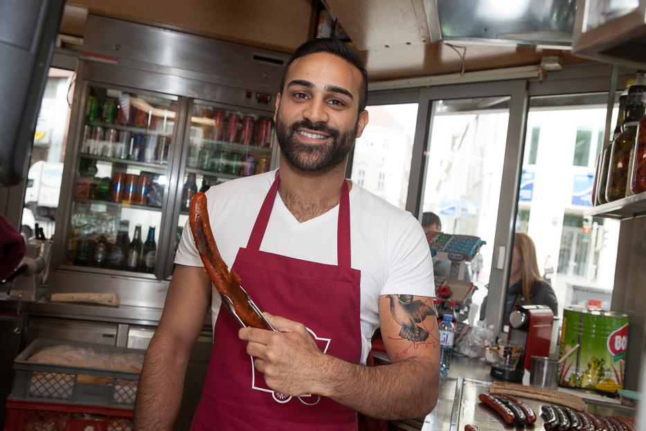 Ein lächelnder Mann mit Tätowierung hält eine Wurst mit einer Zange in einem Imbissstand.
