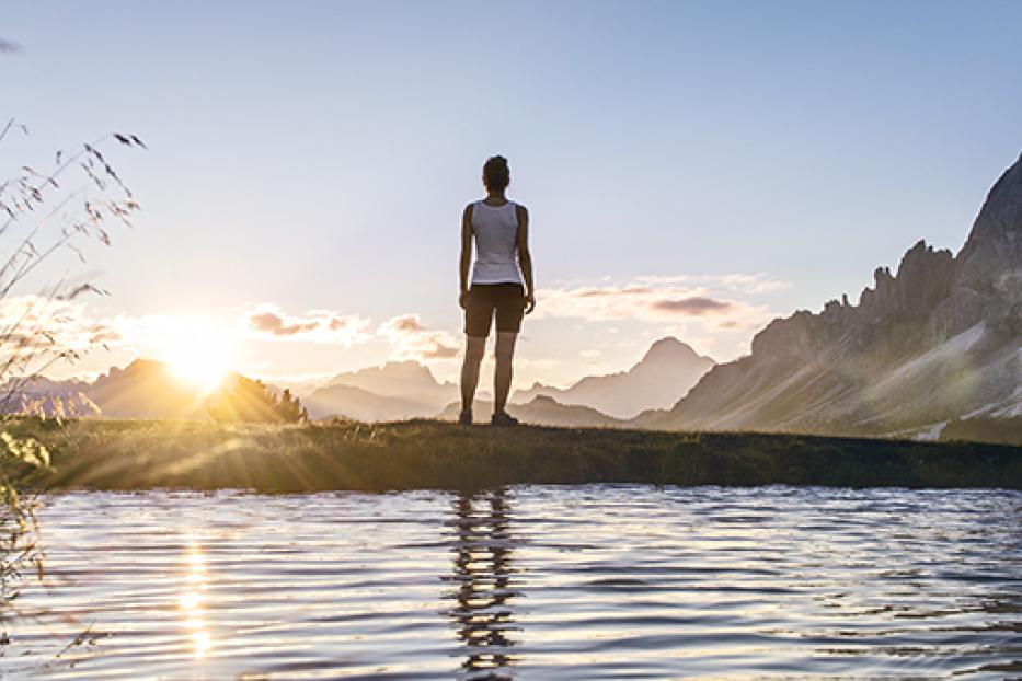 Eine Frau steht am Ufer eines Sees und blickt auf die Berge im goldenen Abendlicht.