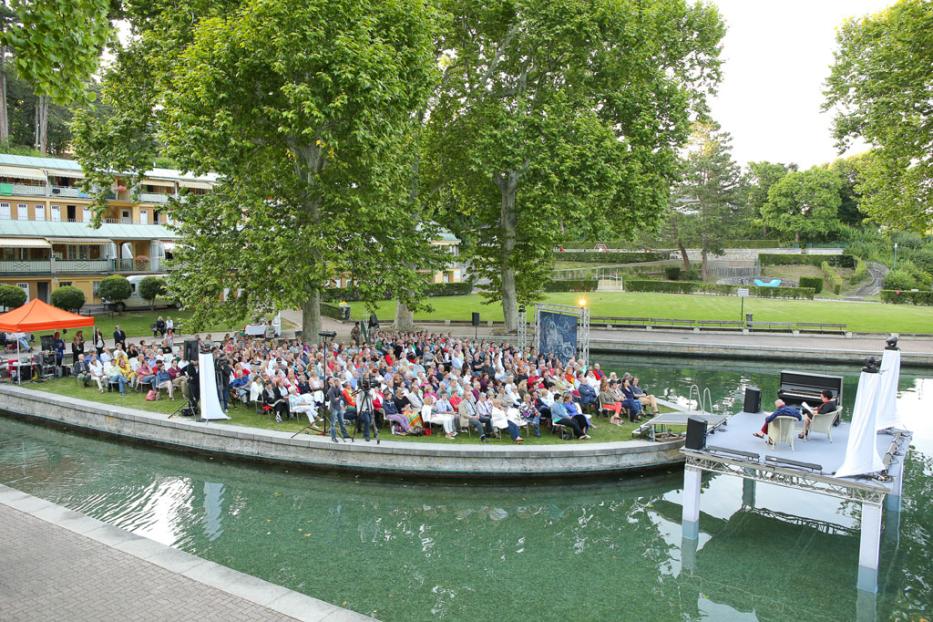 Eine Menschenmenge sitzt auf einer kleinen Halbinsel und schaut einer Aufführung auf einer schwimmenden Bühne zu.