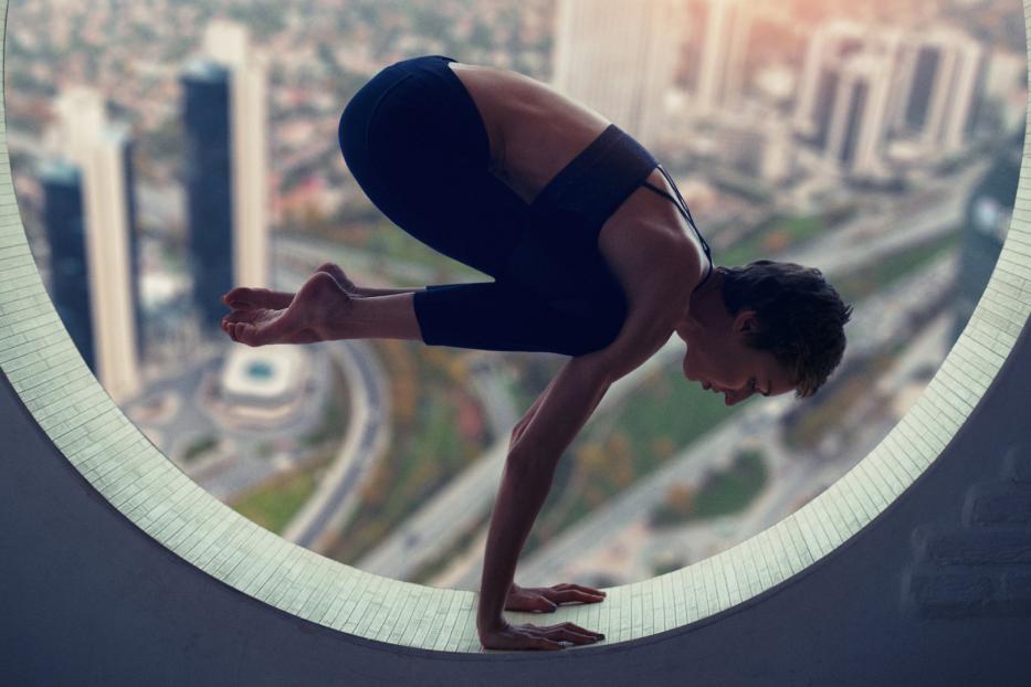 Eine Frau macht Yoga in einer Krähen-Pose vor einem Fenster mit Blick auf die Stadt.