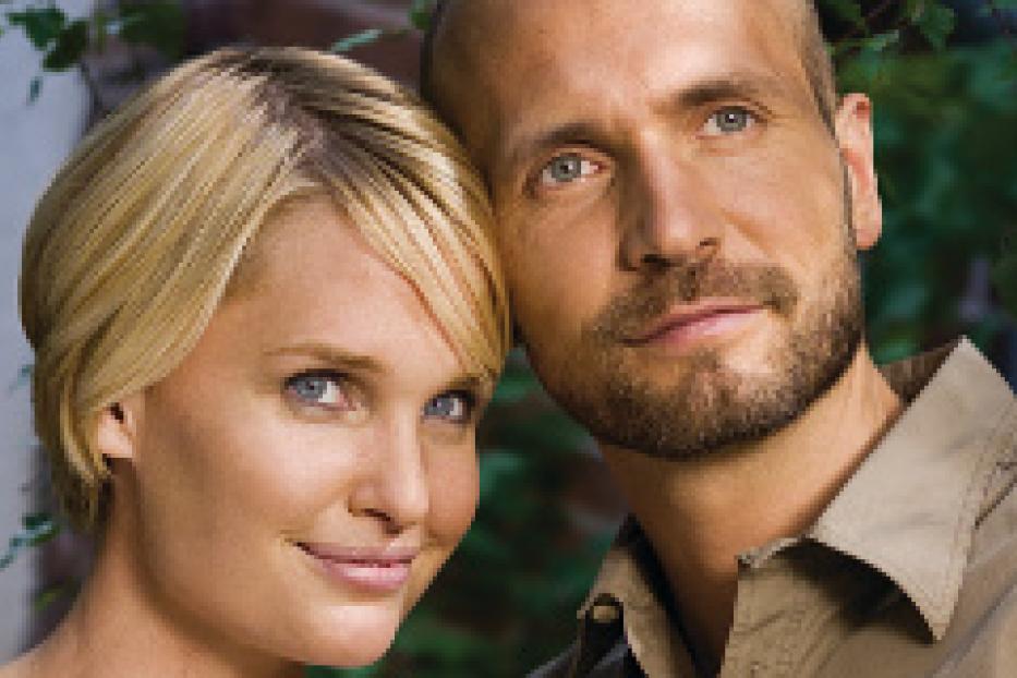 Ein Paar, eine blonde Frau und ein Mann mit Bart, lächeln in die Kamera.