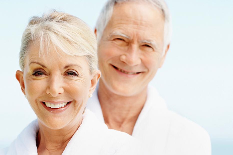 Ein älteres Paar lächelt in Bademänteln vor einem hellen, unscharfen Hintergrund.