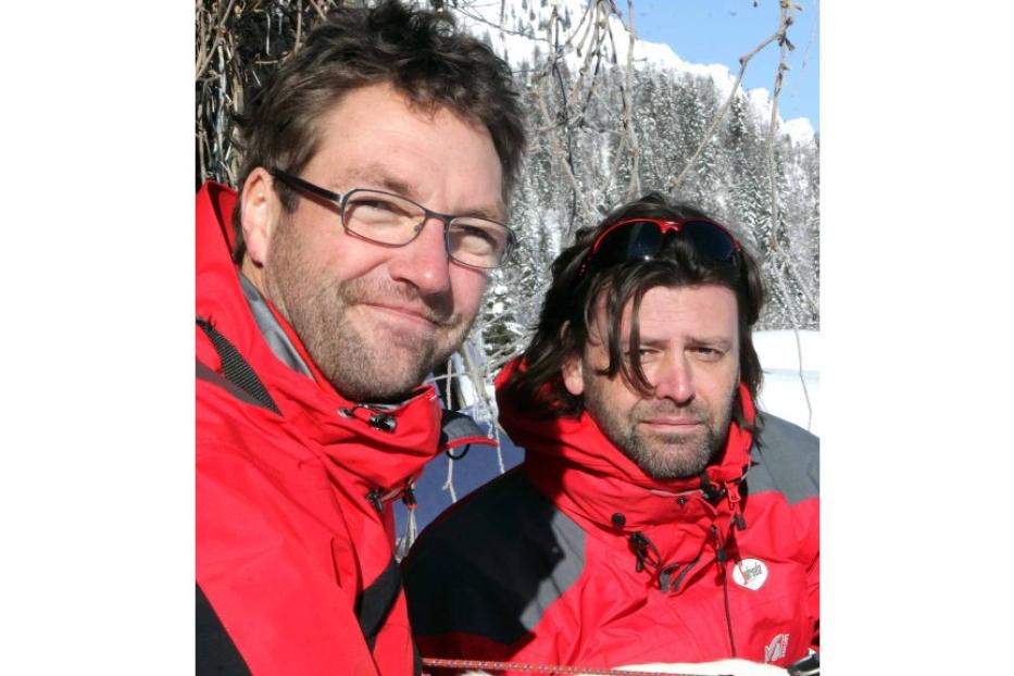 Zwei Männer in roten Jacken posieren vor einer verschneiten Landschaft.