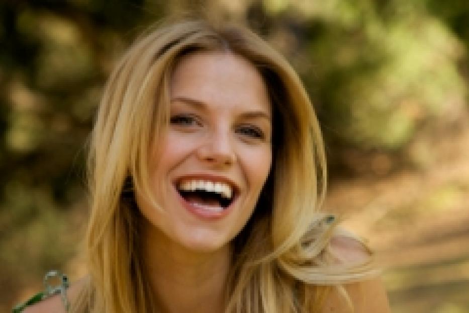 Eine lachende Frau mit blonden Haaren vor einem unscharfen, grünen Hintergrund.