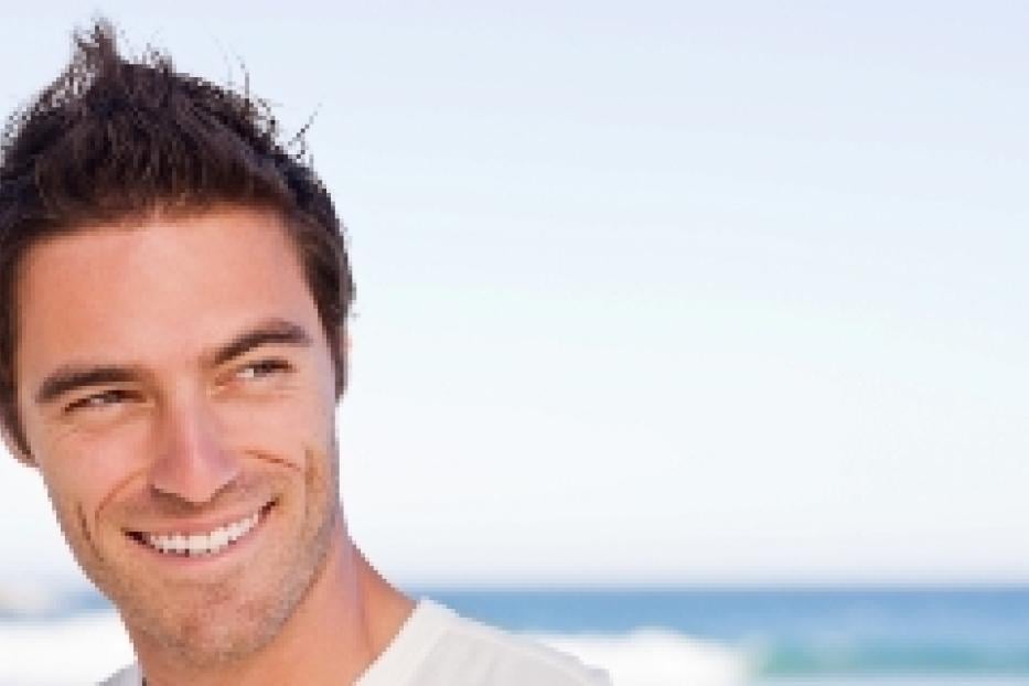 Ein lächelnder Mann mit dunklen Haaren blickt am Strand zur Seite.