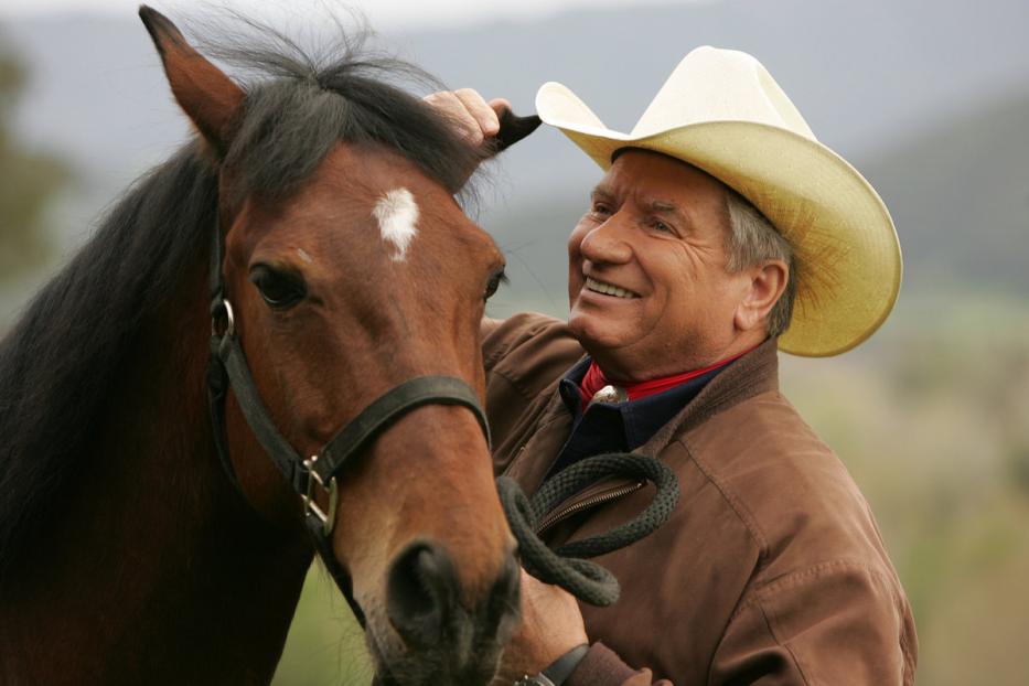 Ein Mann mit Cowboyhut krault ein braunes Pferd hinter dem Ohr.