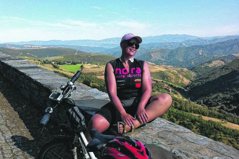 Eine Radfahrerin sitzt auf einer Mauer mit Blick auf eine hügelige Landschaft.