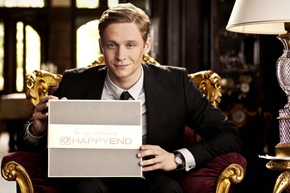 Ein Mann im Anzug sitzt in einem Sessel und hält eine Box mit der Aufschrift „Ihr persönliches Happy End“.