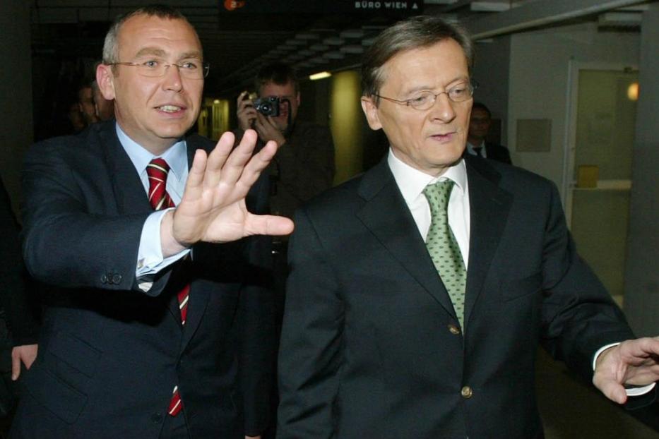 Zwei Männer in Anzügen gestikulieren in einem Flur.