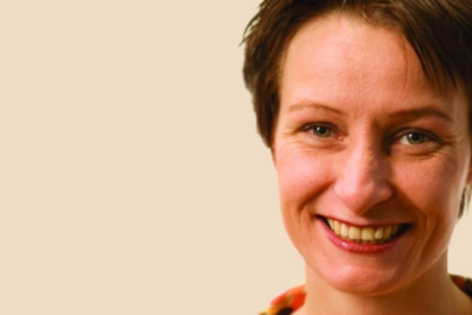 Eine lächelnde Frau mit kurzen braunen Haaren vor einem hellen Hintergrund.