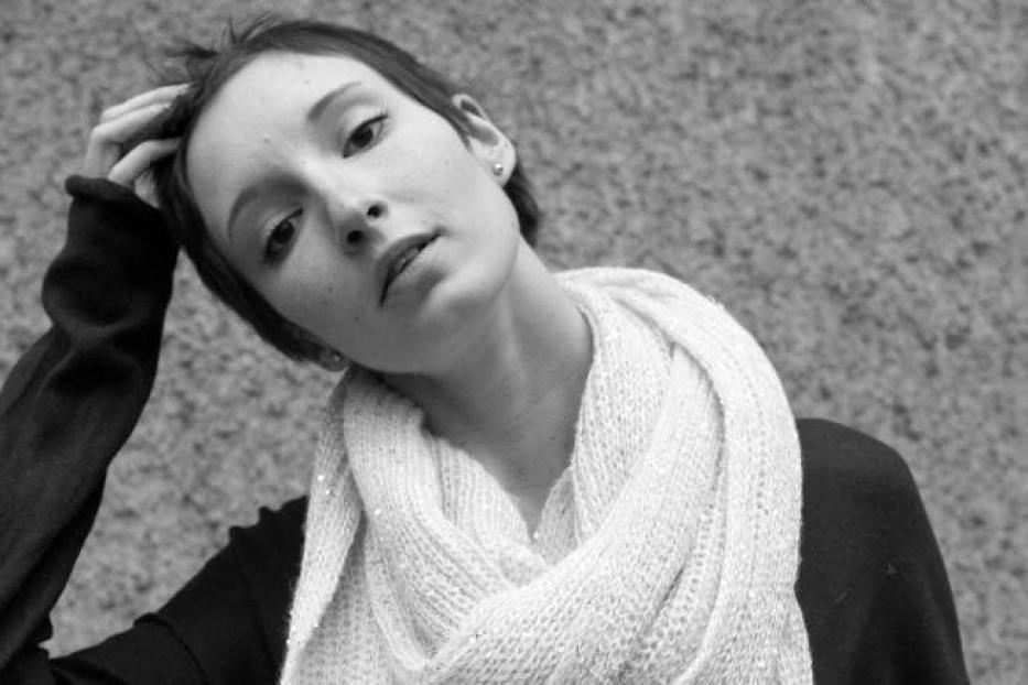 Eine junge Frau mit kurzem Haar und einem weißen Schal vor einer grauen Wand.