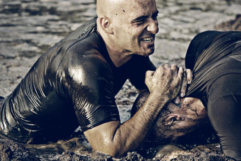 Zwei Männer kämpfen schlammbedeckt im Wasser miteinander.