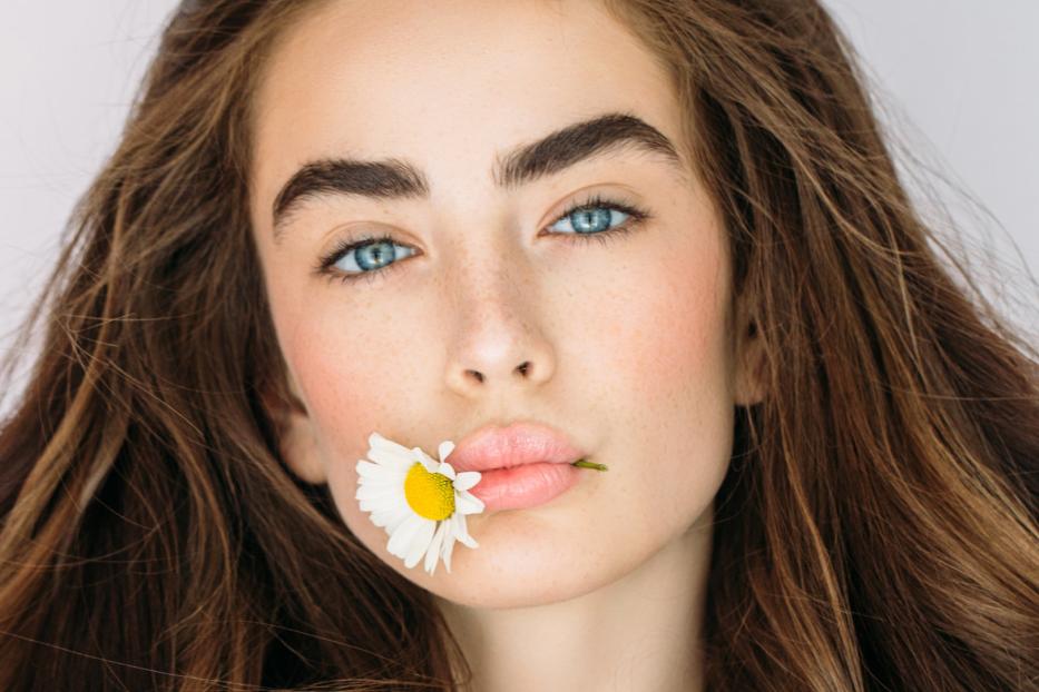 Eine Frau mit braunen Haaren hält eine Gänseblümchenblüte im Mund.