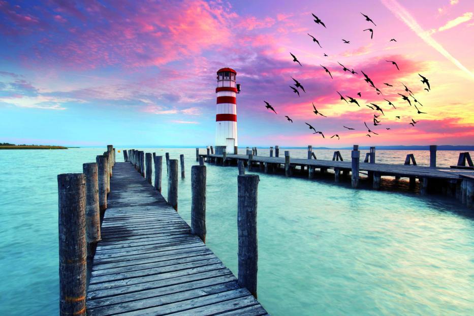 Ein rot-weiß gestreifter Leuchtturm steht am Ende eines Holzstegs bei Sonnenuntergang.