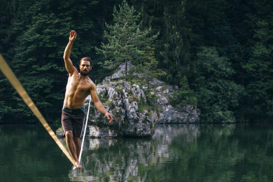 Ein Mann balanciert barfuß auf einer Slackline über einem See vor bewaldeter Kulisse.