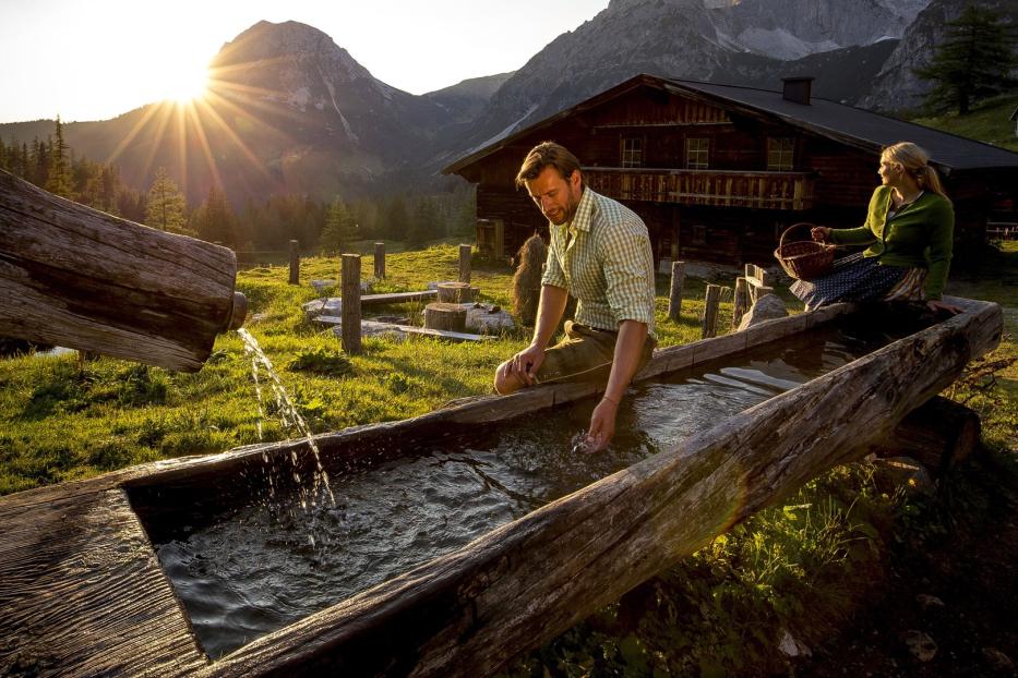 Ein Mann und eine Frau sitzen an einem hölzernen Brunnen vor einer Berghütte.