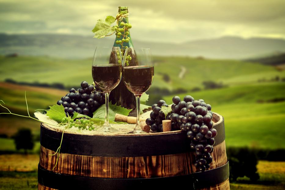 Auf einem Weinfass stehen zwei Gläser Rotwein und Trauben vor einer Hügellandschaft.