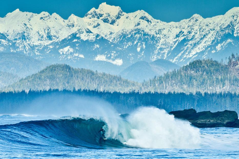 Ein Surfer reitet auf einer Welle vor einer Bergkulisse mit schneebedeckten Gipfeln.