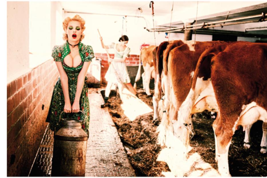 Eine Frau im Dirndl steht mit einem Milcheimer in einem Kuhstall.