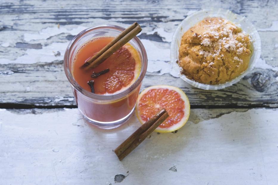 Ein Glas mit einem Getränk, einer Orangenscheibe, Zimtstangen und einem Muffin auf einem Tisch.