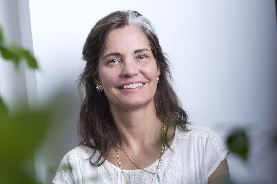 Eine lächelnde Frau mit braun-grauem Haar und weißer Bluse, umgeben von grünen Blättern.