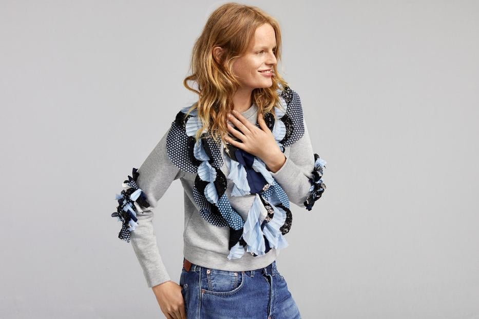 Eine Frau mit blonden Haaren trägt einen grauen Pullover und eine Halskette mit Rüschen.