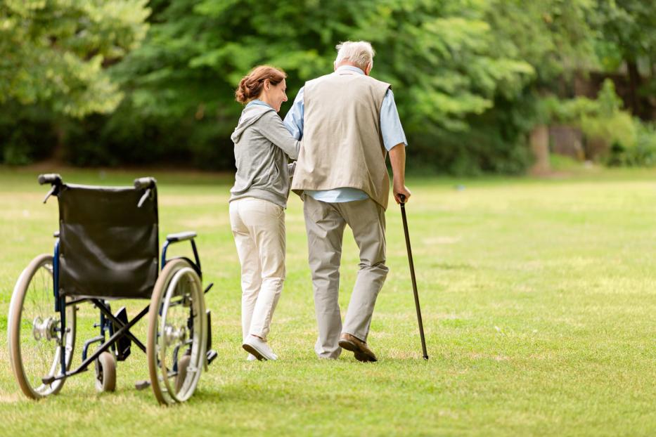 Eine Frau unterstützt einen älteren Mann mit Gehstock bei einem Spaziergang im Park. Im Vordergrund steht ein Rollstuhl.