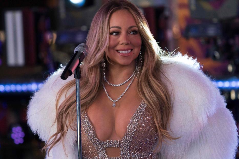 Mariah Carey steht lächelnd mit Glitzerkleid und weißem Fellmantel vor einem Mikrofonständer