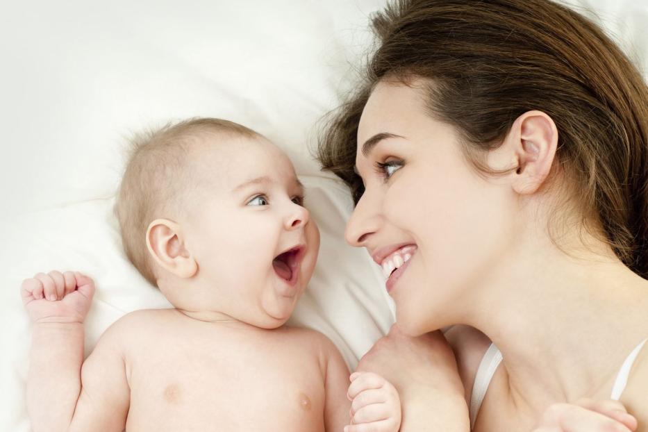 Eine Mutter lächelt ihr lachendes Baby an, während sie auf einem weißen Laken liegen.