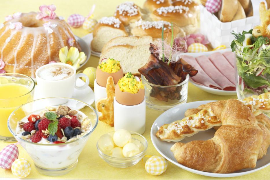 Ein reichhaltiges Osterfrühstück mit Kuchen, Croissants, Eiern, Müsli und Aufschnitt auf einem gelben Tisch.
