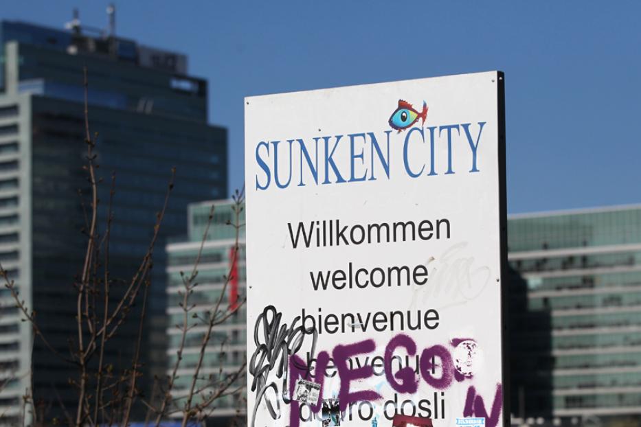 Ein Schild mit der Aufschrift „Sunken City“ in mehreren Sprachen vor modernen Gebäuden.