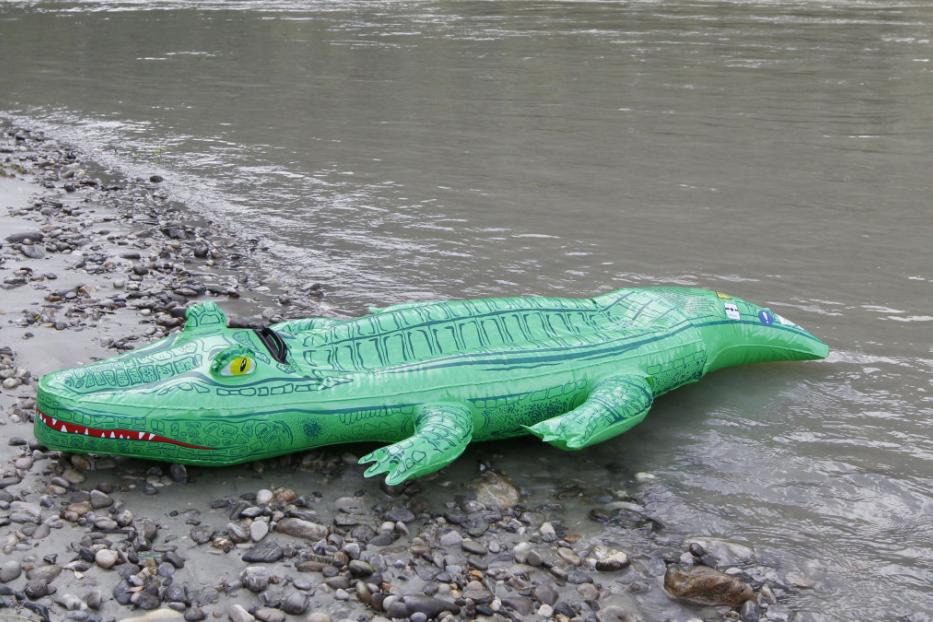 Ein aufblasbares, grünes Krokodil liegt am Ufer eines Flusses.
