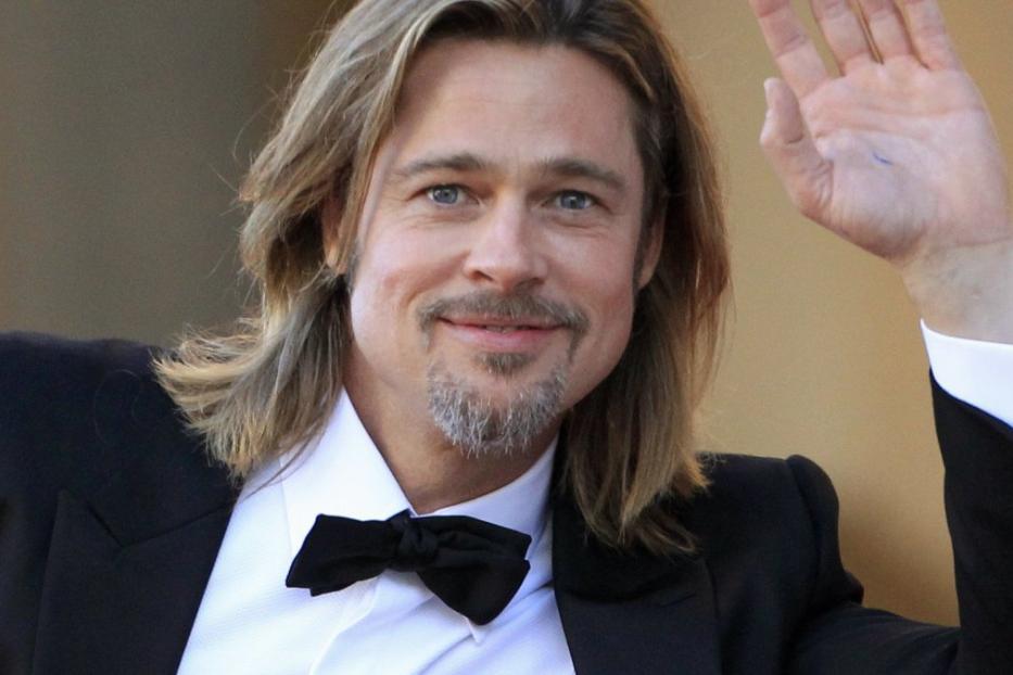 Brad Pitt lächelt und winkt in einem schwarzen Anzug.