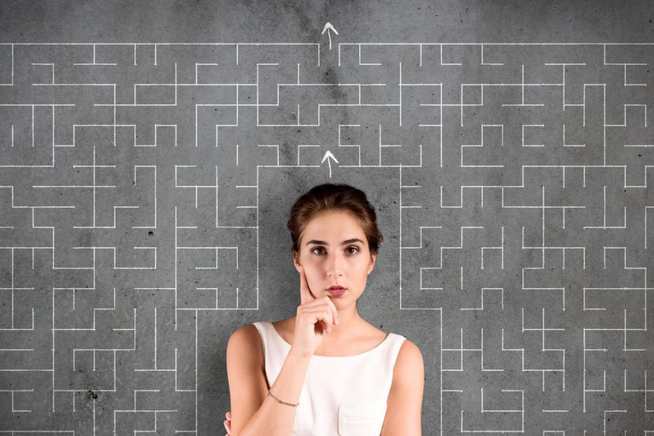 Eine Frau steht vor einer grauen Wand mit einem aufgemalten Labyrinth und Pfeilen.