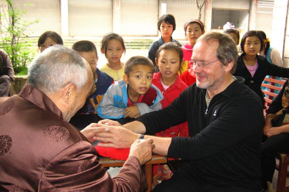Ein älterer Mann untersucht die Hand eines anderen Mannes, während Kinder zusehen.