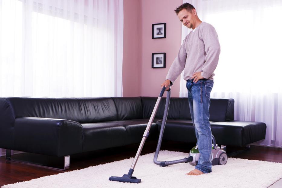 Ein Mann staubsaugt barfuß einen weißen Teppich in einem Wohnzimmer.