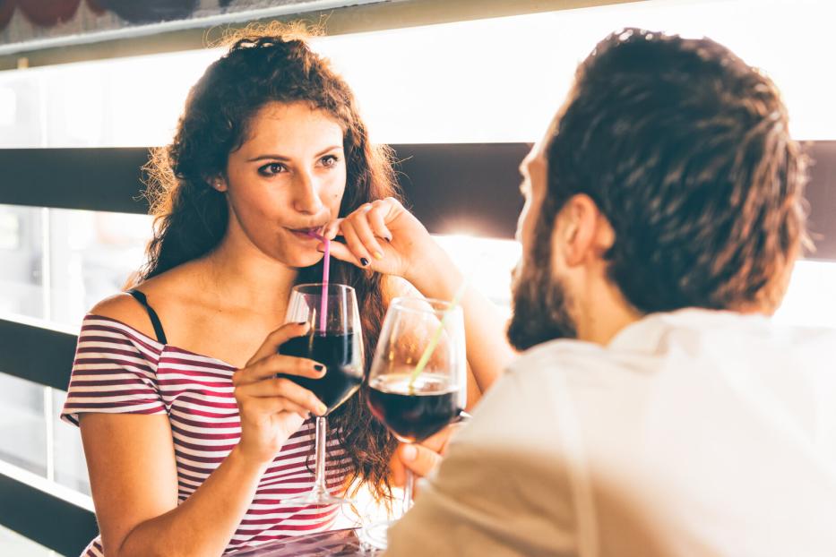 Eine Frau mit lockigem Haar und ein bärtiger Mann sitzen sich gegenüber und trinken ein Getränk aus Gläsern mit Strohhalmen.