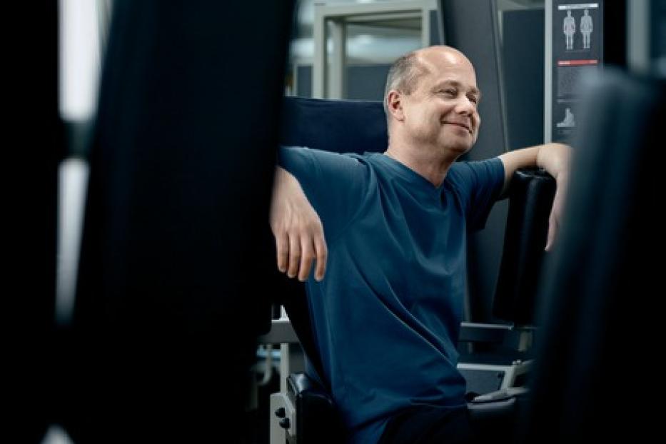 Ein Mann mittleren Alters sitzt entspannt in einer Trainingsmaschine im Fitnessstudio.