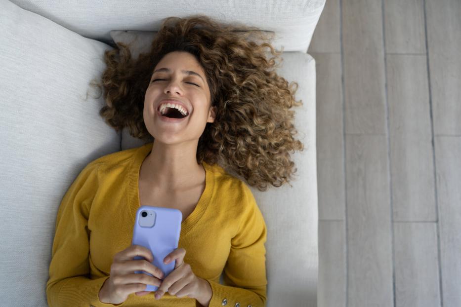 Eine lachende Frau liegt mit einem Handy auf einem Sofa.
