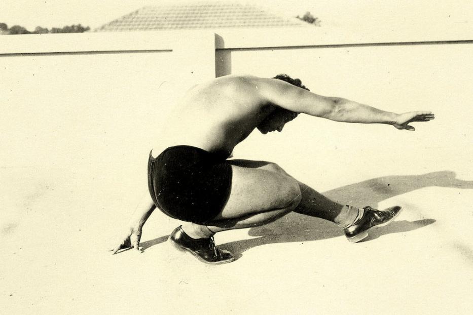 Ein Mann in Badehose und Schuhen kauert mit ausgestreckten Armen auf dem Boden.