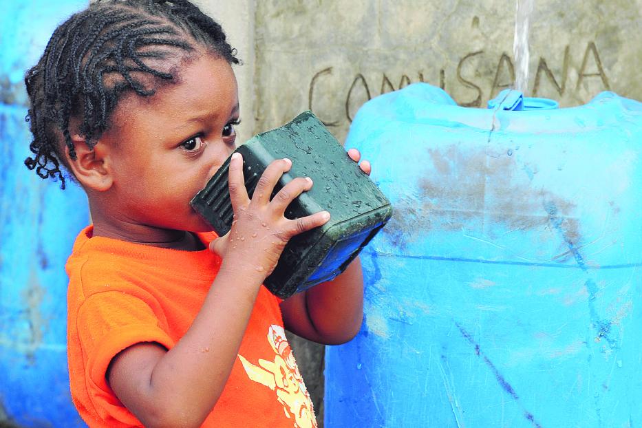 Ein kleines Mädchen trinkt Wasser aus einem Becher vor einem Hilfswerk-Brunnen.