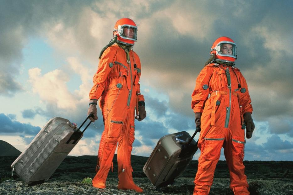 Zwei Personen in orangefarbenen Raumanzügen ziehen Koffer über eine felsige Landschaft.