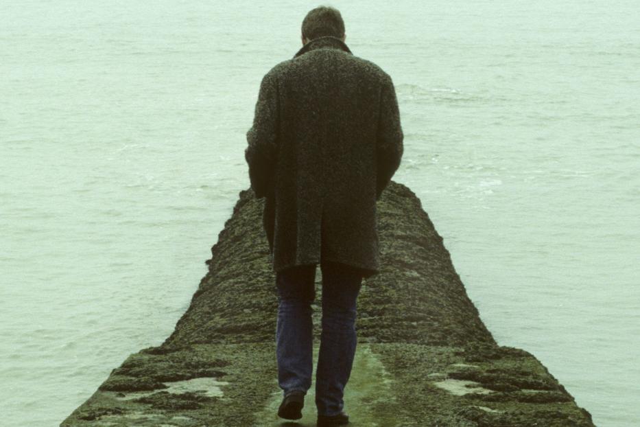 Ein Mann in Mantel geht auf einem Steg ins Meer.