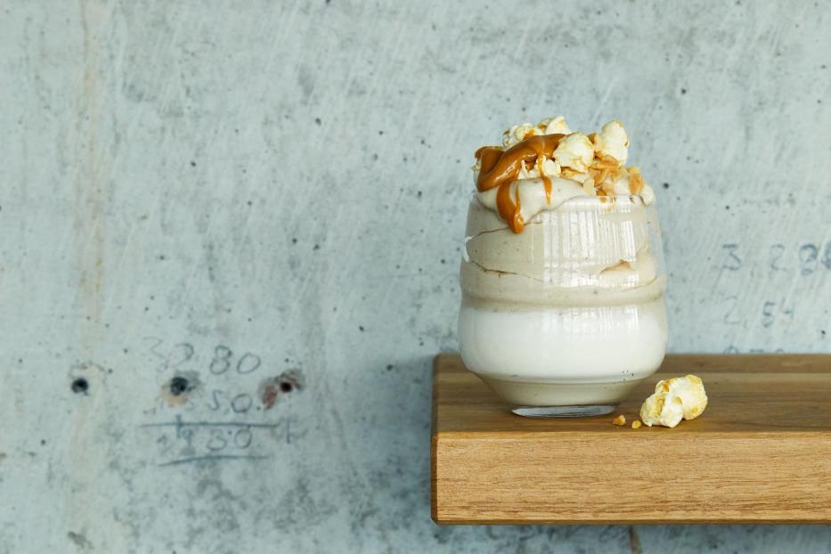 Ein Glas Dessert mit Karamell, Popcorn und einer hellen Creme steht auf einem Holzbrett.