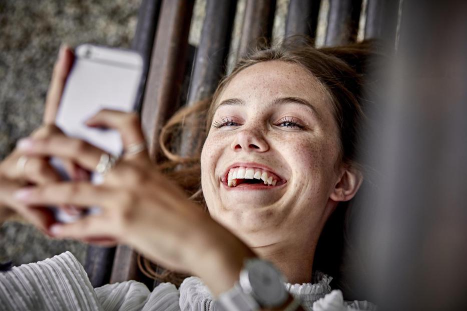 Laughing young woman lying on a bench using cell phone - Stock-Fotografie